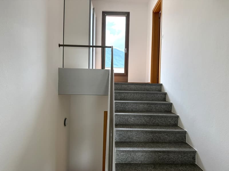 The image shows a staircase with gray granite steps leading up to a window with a view of the mountains outside. The staircase has a metal railing and there is a white wall on one side.