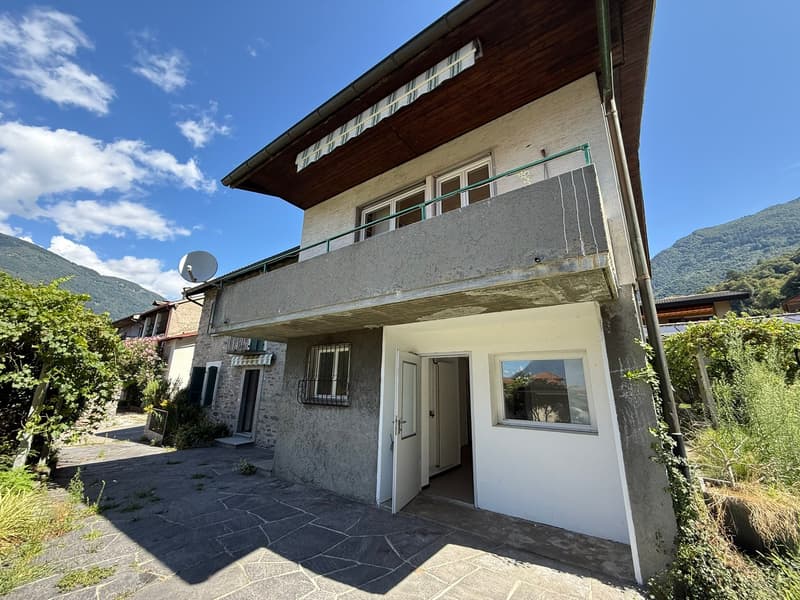 2-story house with wooden roof, stucco exterior, balcony, and paved parking area. The house is surrounded by greenery and mountains in the background.
