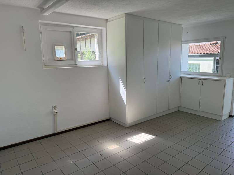 The image shows a room with white walls, a tiled floor, and a large built-in wardrobe along one wall. There is a window that provides natural light to the space. The room appears to be a bedroom or living area.