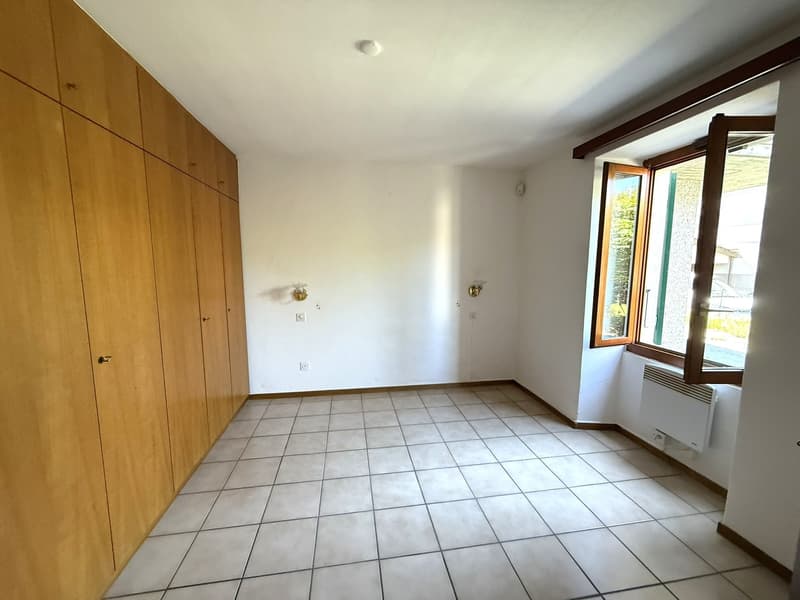 The image shows a spacious bedroom with wooden built-in wardrobes along one wall. The room has a tiled floor and a large window that provides natural light. The walls are painted in a neutral color, and the room appears to be clean and well-maintained.