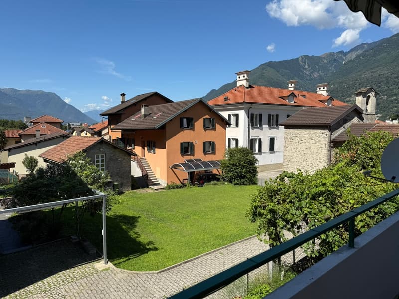 Multi-story residential buildings with red tile roofs, surrounded by mountains, with a grassy lawn and paved walkway in the foreground.