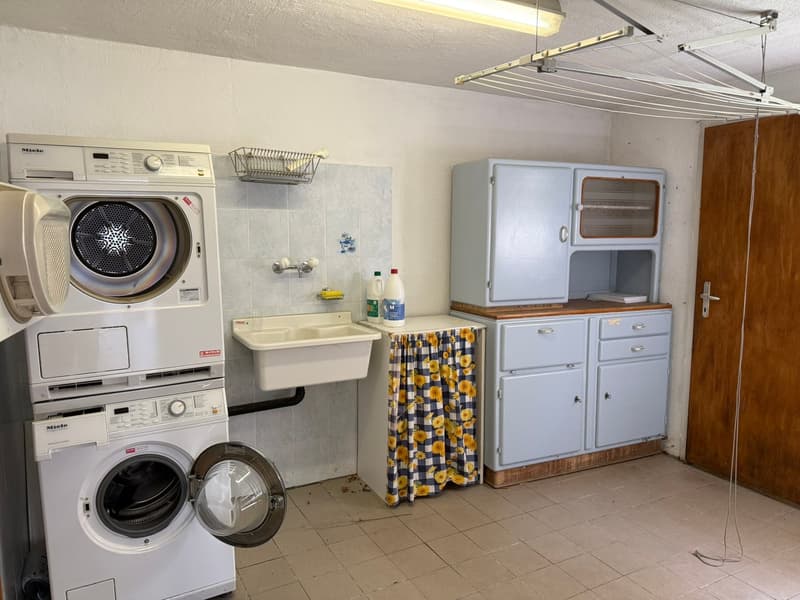 The image shows a laundry room with a Miele washing machine and dryer, a sink, a curtain-covered storage unit, and a refrigerator. The room has tile flooring and appears to be a functional laundry space.