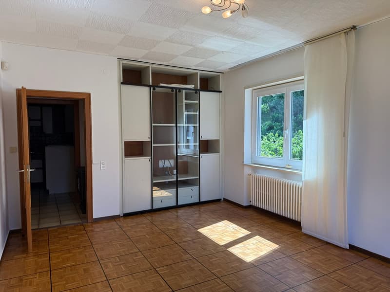 The image shows a bedroom with a large built-in wardrobe along one wall. The room has a tiled floor and a window overlooking some greenery outside. The room appears to be well-lit with natural light coming in through the window.