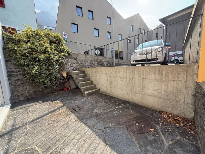 Paved courtyard with a car parked, concrete retaining wall with stairs, fence, metal grates on the floor, bushes, and some objects leaning on the wall.