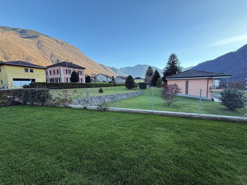 well-kept lawn, surrounded by a fence, multiple buildings in the background, a slope with houses and trees, a clear view of the mountains