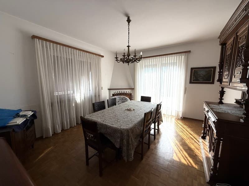 A dining area with a long table, a chandelier, wooden chairs, white curtains, and a fireplace