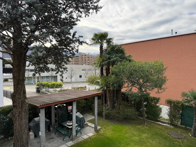 Outdoor covered area with metal roof, green bench, table, and a gas tank, surrounded by green grass, trees, and bushes.