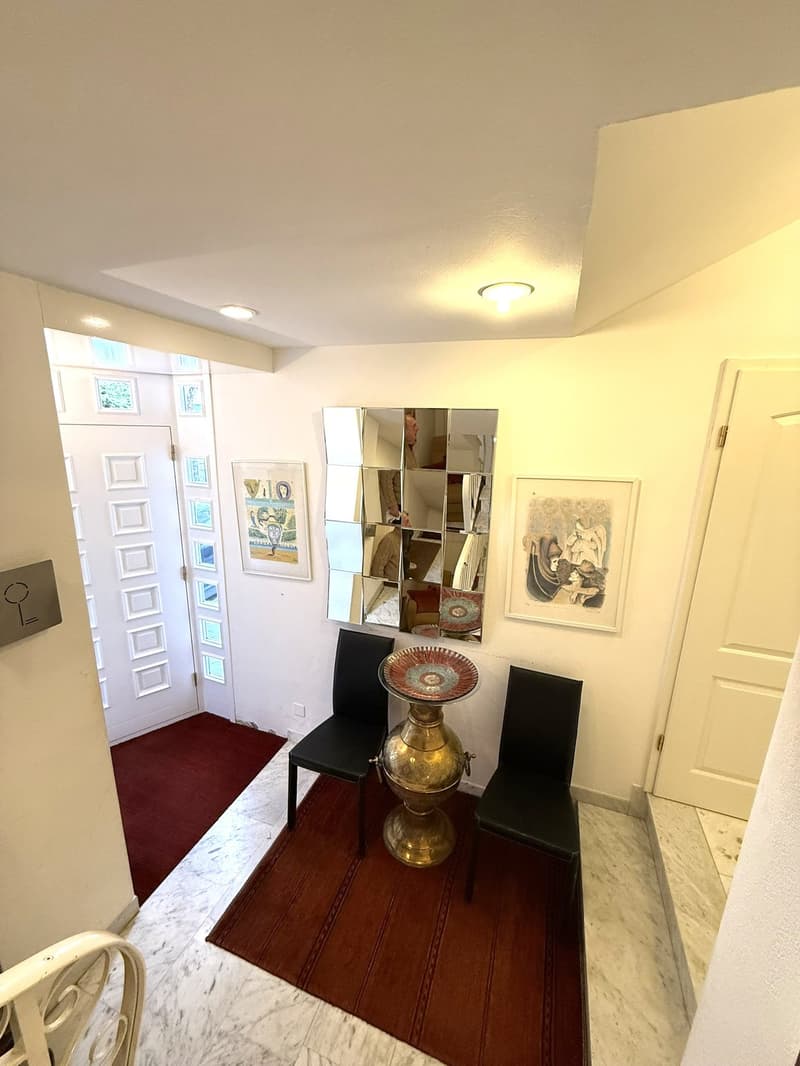 A hallway with a red carpet, two black chairs, a golden table, a large mirror, and two framed art pieces on the wall.