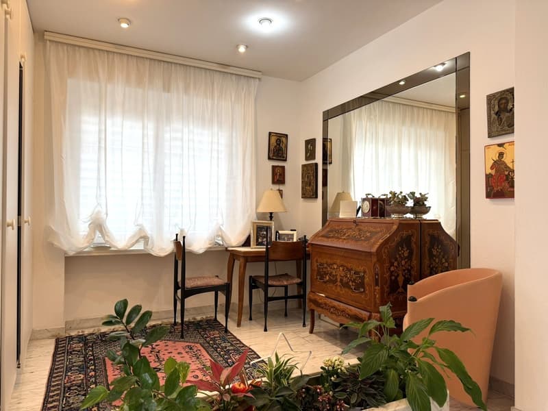 A room with a desk, chairs, a mirror, a rug, and plants. The room has a window with white curtains. The desk has a lamp, a picture frame, and a clock. The mirror is mounted on the wall, and there are picture frames on the wall. The room has a wooden floor