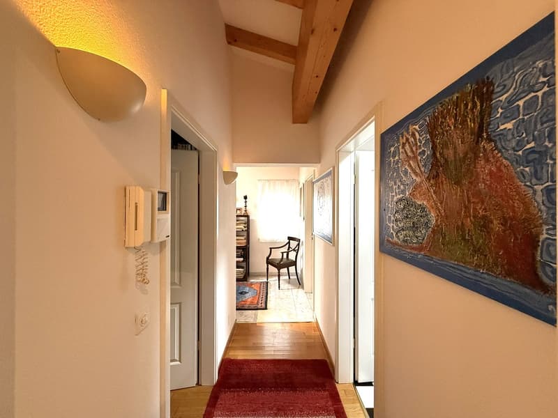 Long hallway with wooden flooring, paintings on the wall, telephone, hallway lamp, and rug.