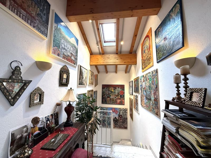 wooden ceiling, wall with various artwork and paintings, desk with red cloth, lamp, books on shelf, small gate at the end