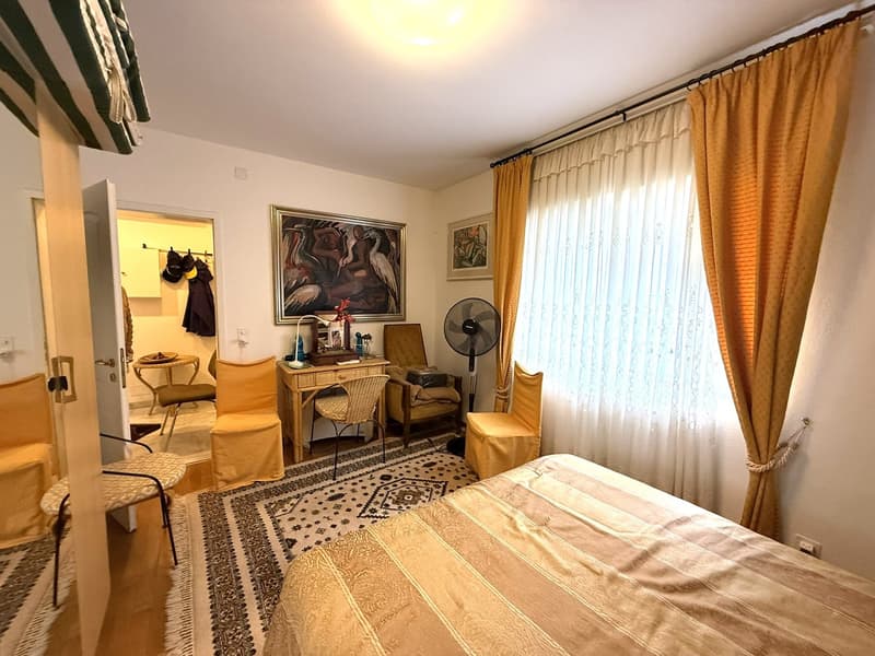 a large bed, desk, table, chairs, paintings, rug, curtain, fan