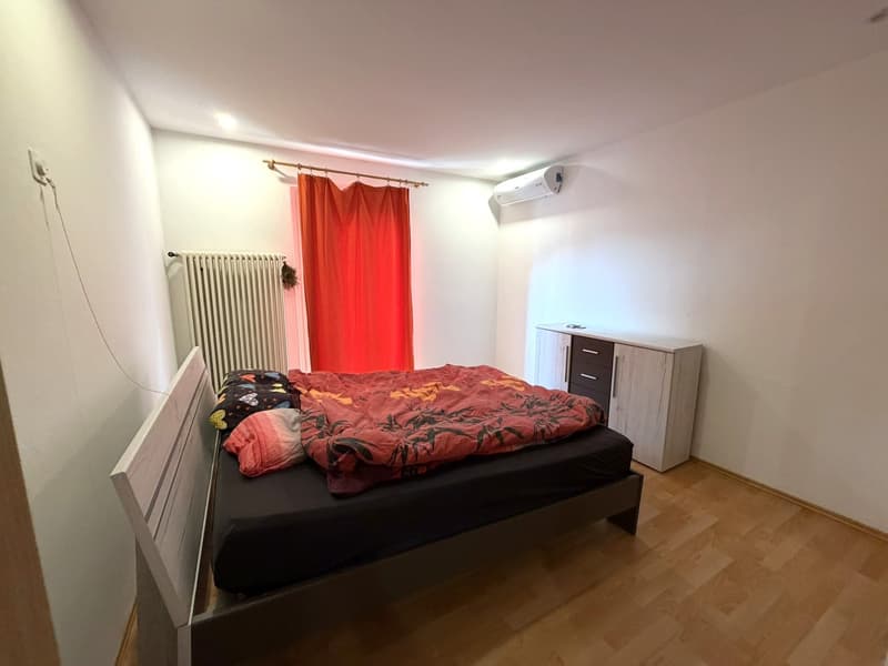 Bedroom with a bed, red blanket, pillows, white walls, wooden floor, white cabinet, red curtain, white radiator, air conditioner