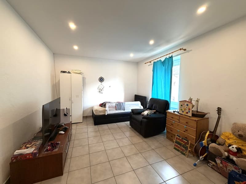 Living room with a flat screen TV, a wooden cabinet, a black chair, a bed, a wooden cabinet with drawers, a guitar, a teddy bear, and a toy boat.