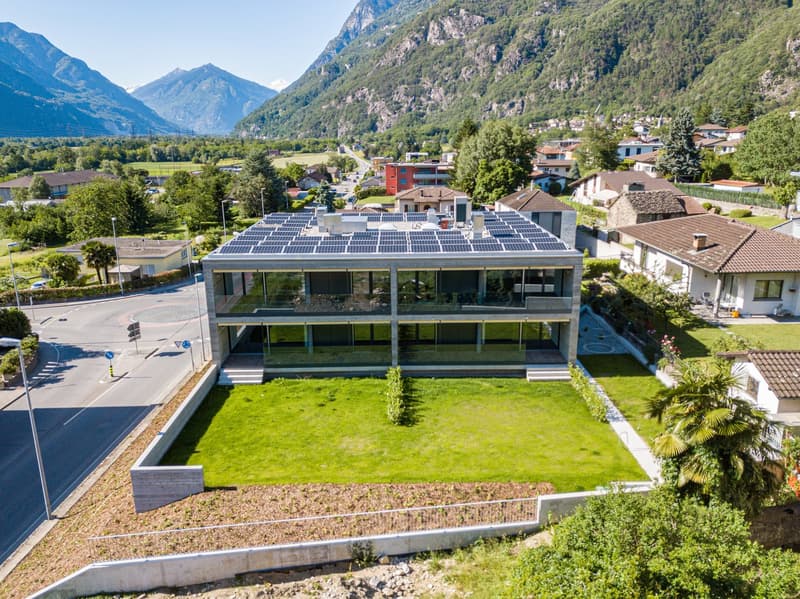 Modern house with solar panels on the roof, glass facade, surrounded by greenery, mountains in the background