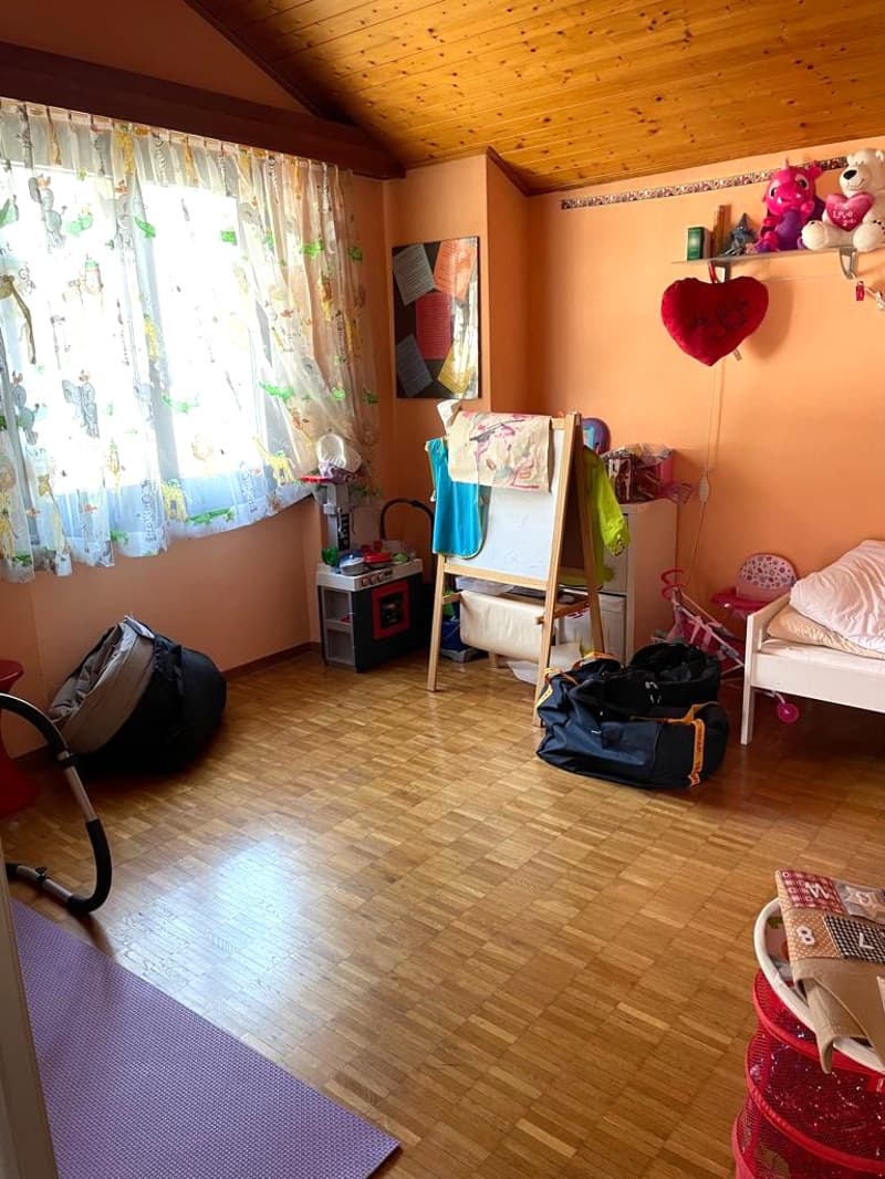 Wooden floor, pink walls, large window with curtains, toys and bags on the floor, small table with a toy kitchen, clothes hanging on a wooden rack, a small bed with a pillow, a shelf with stuffed animals and a heart