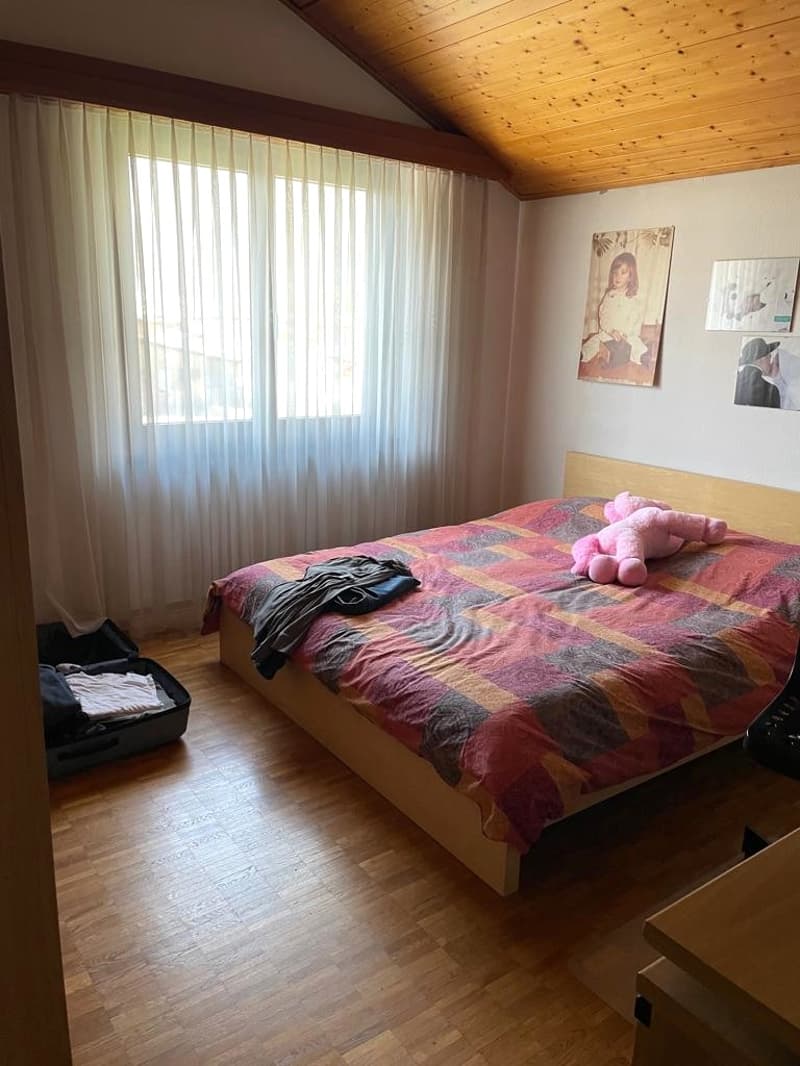 Bedroom with wooden floor, bed, window with white curtains, wooden ceiling, two picture frames on the wall, pink stuffed toy on the bed, suitcase on the floor.