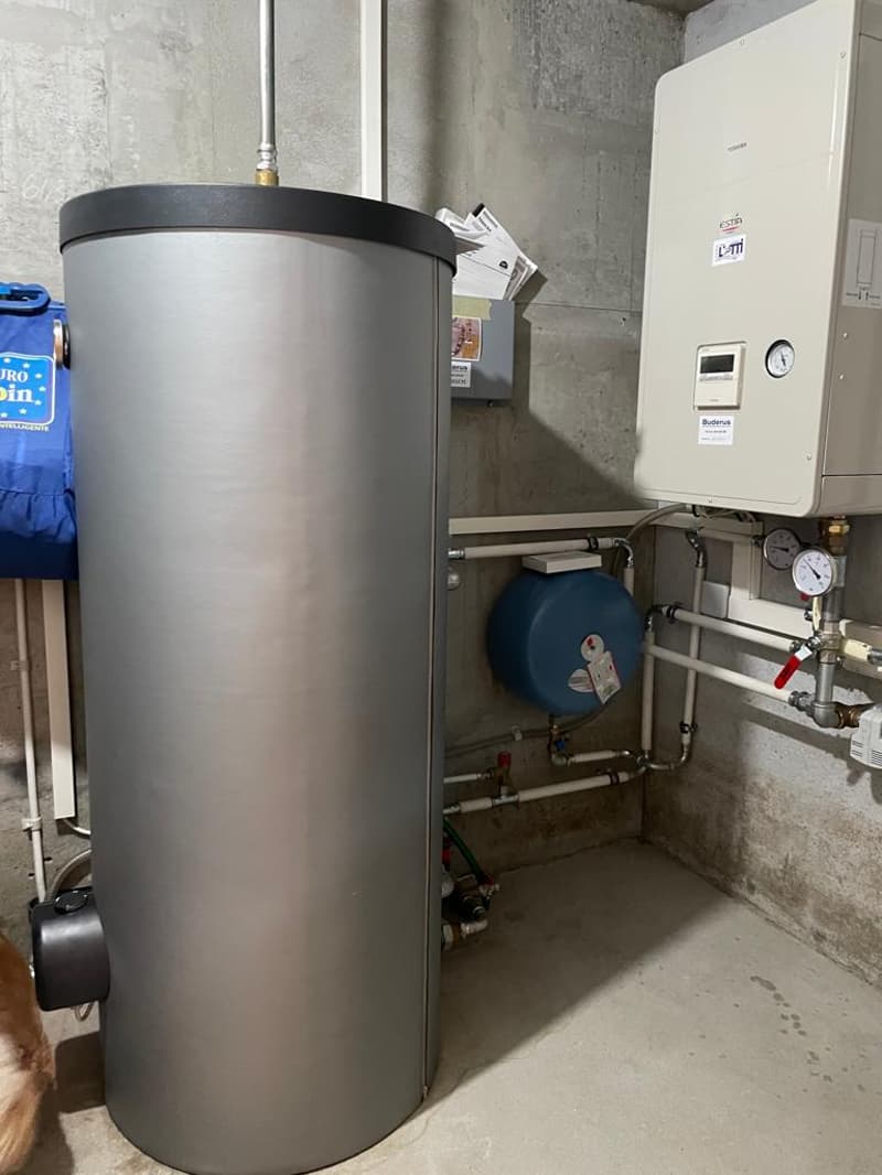 A room with a large silver water heater, a blue water tank, and a white water heater with a digital display. There are also several pipes and gauges attached to the wall.