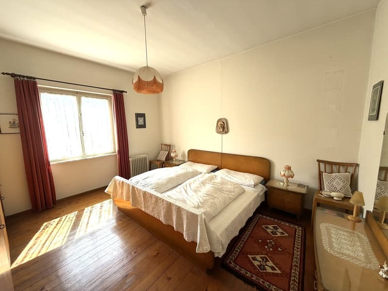 Bedroom with wooden floor, large bed, white sheets, pillows, bedside tables, lamps, radiator, window with red curtains, wall decorations, rug, wooden chair, mirror