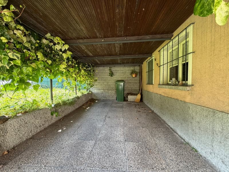 covered walkway with stone floor, wooden ceiling, green trash bin, broom, plants, and a window with bars
