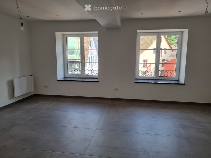 Empty room with white walls, gray tiles, two windows, power outlets, window sills, ceiling light fixture.
