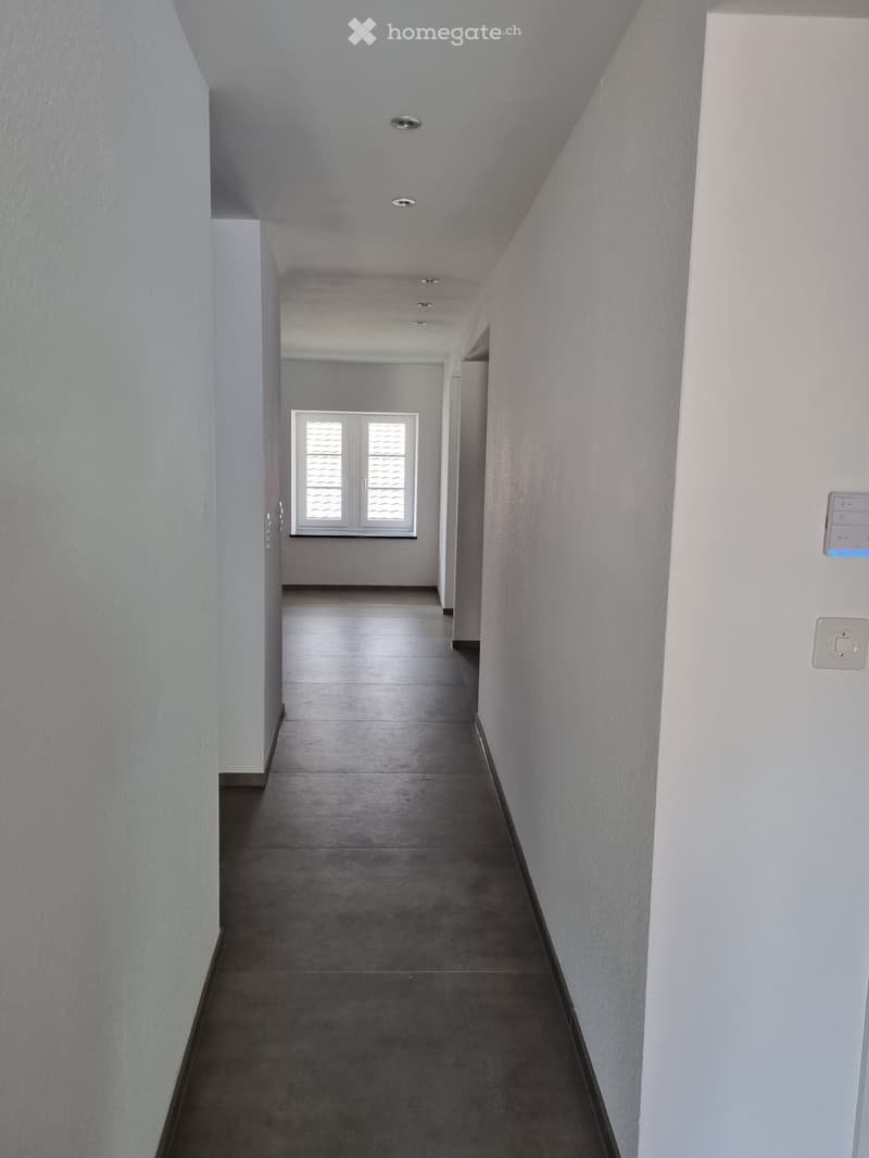 White walls, gray floor, white ceiling with recessed lighting, small window on the left wall, white door on the right wall