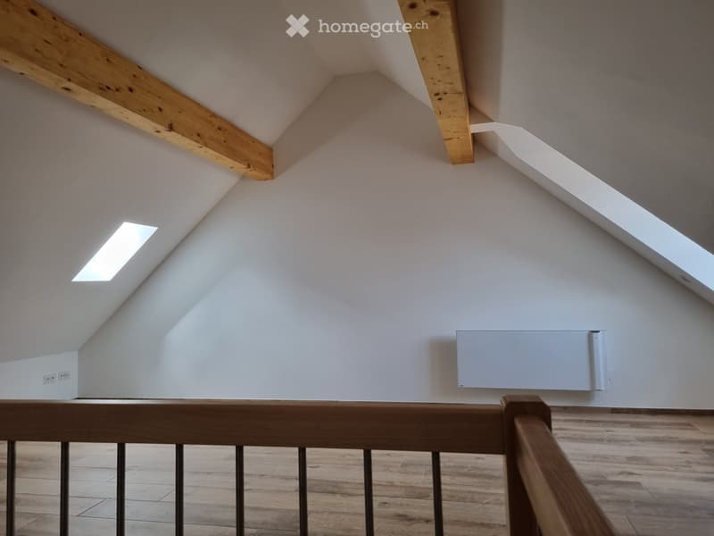 Open attic space with wooden beams, white walls, wood flooring, small skylight, and mounted radiator