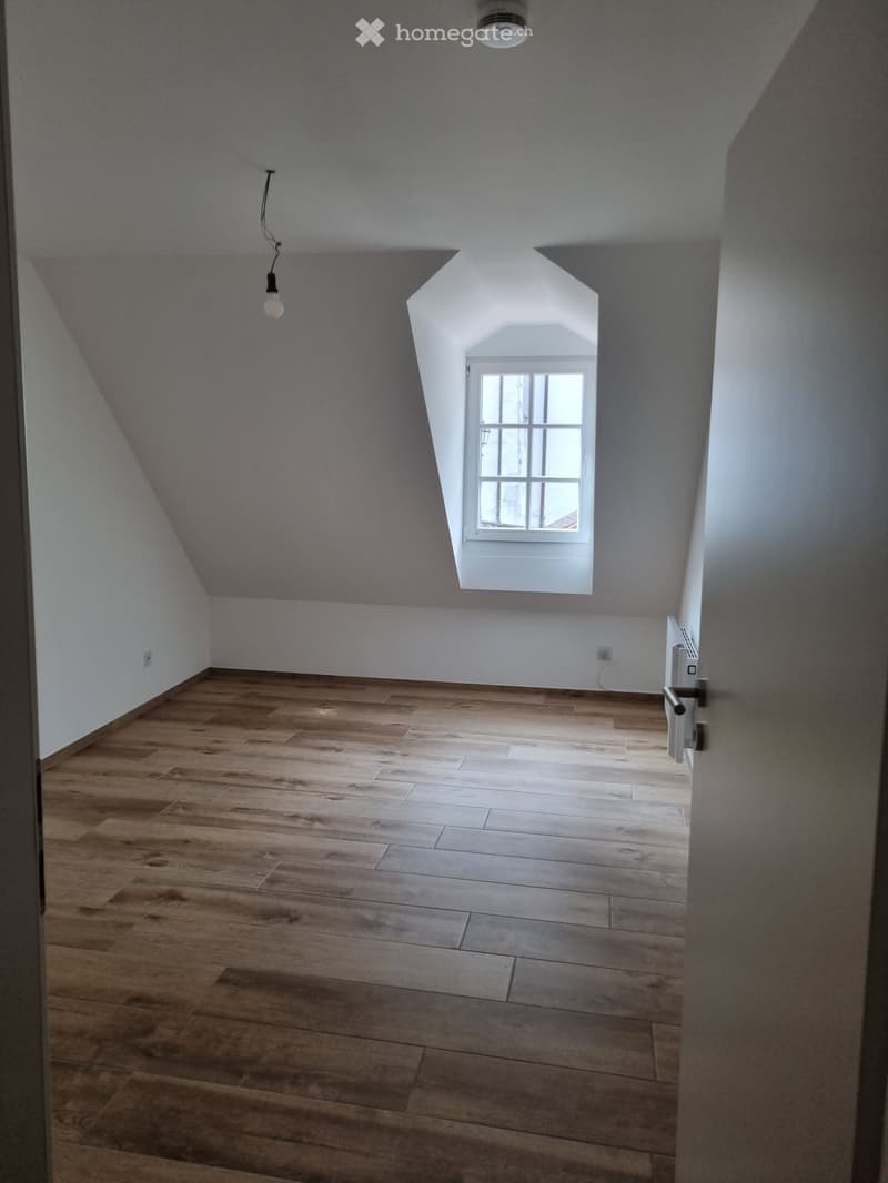 empty attic room with a window, white walls, wooden floor, an outlet and a lamp hanging from the ceiling