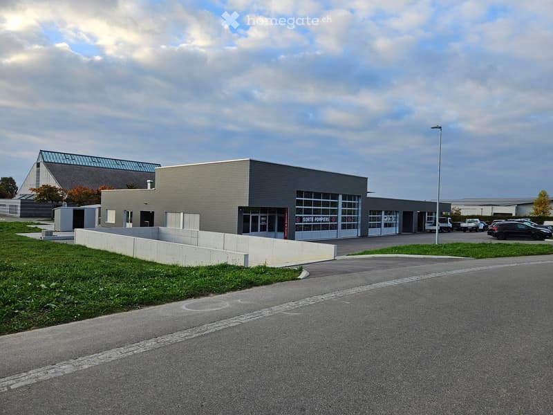 A grey building with a glass door and several windows, parking lot, cars parked in front of it, grassy area in front, street light