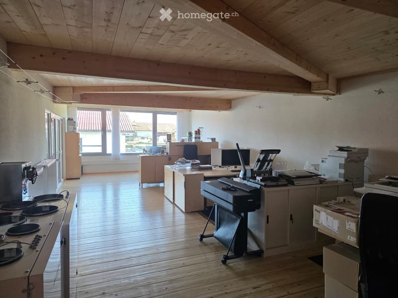 Empty office room with wooden floor, wooden beam ceiling, large windows, white walls, cabinets, computer monitors, printers, various office equipment, small office furniture, boxes