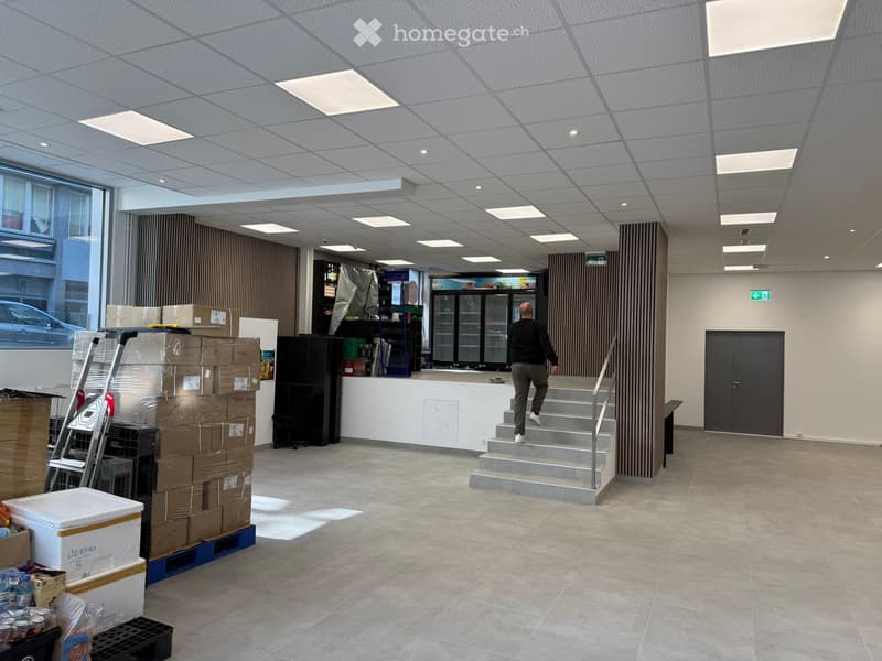 Empty room with tiled floor, white walls, ceiling with recessed lighting, a man walking up the stairs, a ladder, boxes, and a refrigerator.