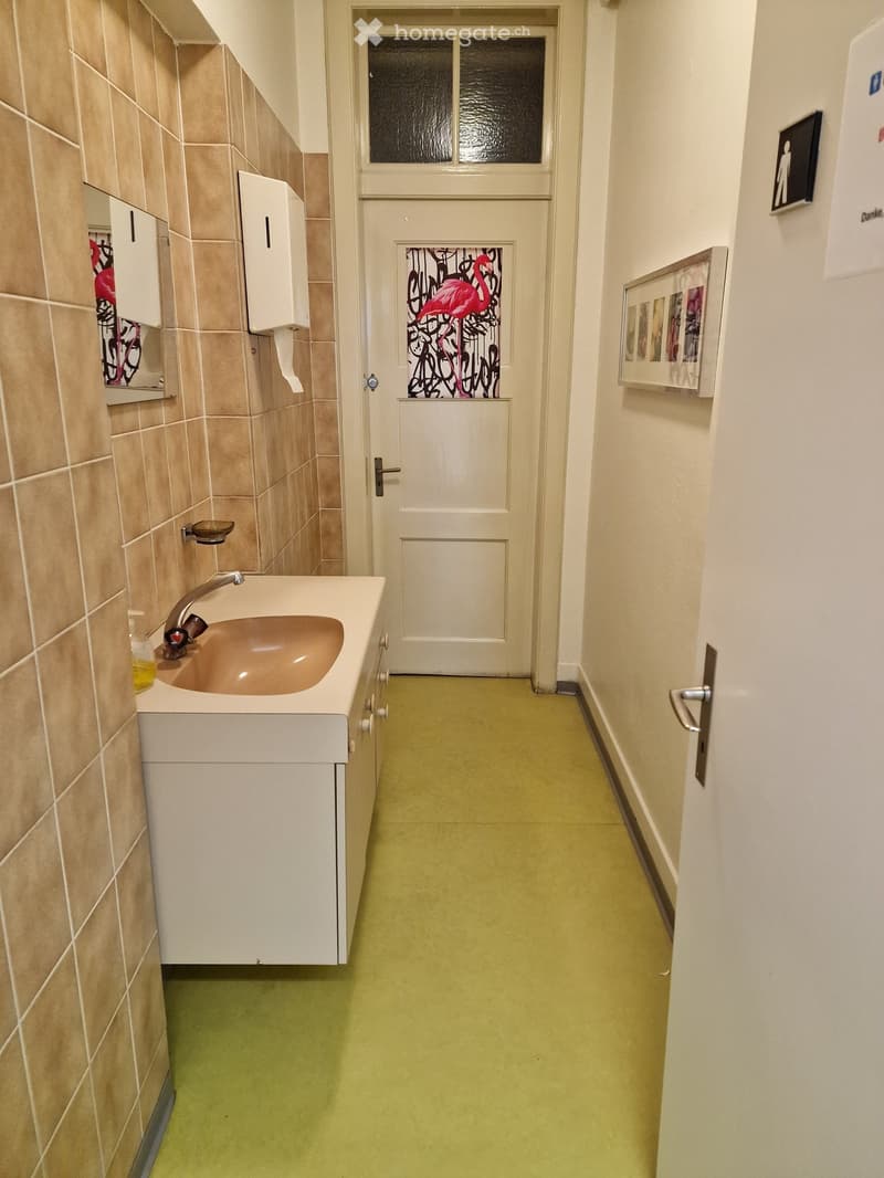 A hallway leading to a bathroom with white cabinets, a sink with a faucet, beige tiles on the wall, and a framed artwork on the wall.