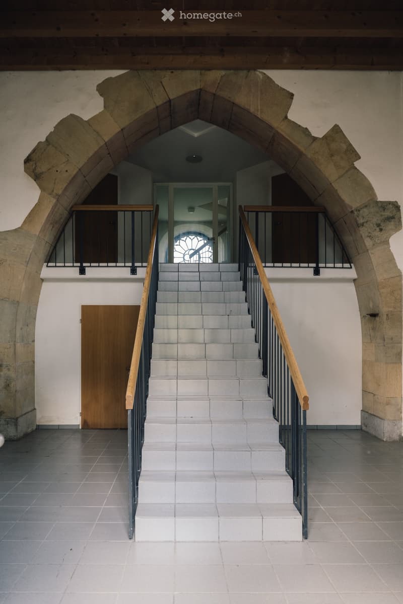 Stone building, arched entrance, white tiled floor, staircase with wooden handrails, metal railings on the upper floor