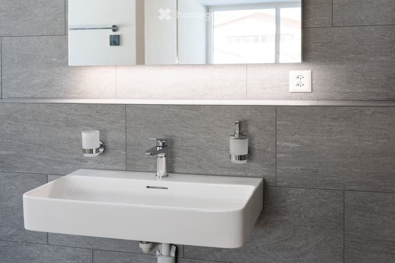 white sink, faucet, mirror, soap dispenser, power outlet, gray tiles