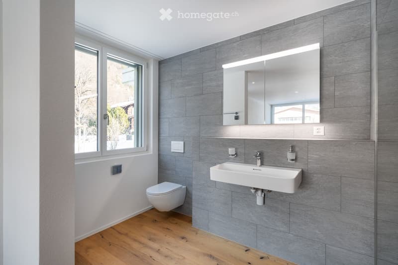 A modern, minimalist bathroom with a wall-mounted toilet, large mirror, silver fixtures, and wooden floor. Natural light comes through a window.