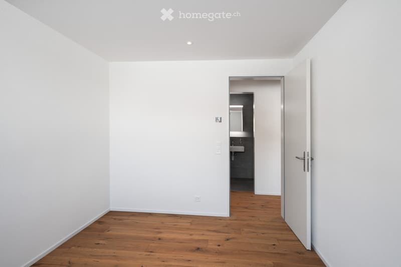 Empty room with wooden floor, white walls, open door leading to bathroom with sink.