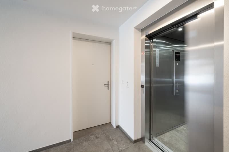 An elevator in a minimalistic corridor, with a white wall, and a white door with a metal handle.