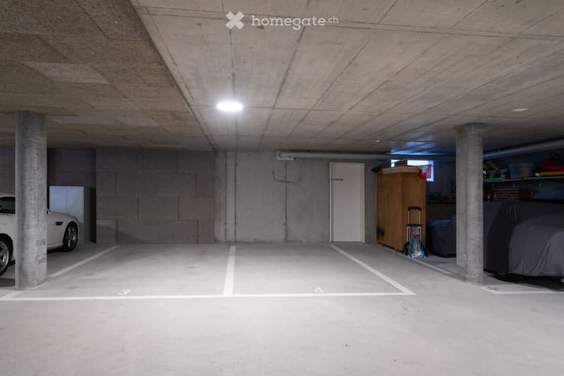 Underground parking lot, concrete walls, gray floor, two columns, ceiling lights, storage area, parked white car, storage boxes