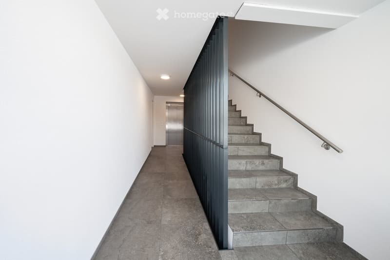 A long narrow hallway with white walls and tile flooring, featuring a metal handrail on one side leading to a staircase, an elevator on the opposite side, and a small room at the end.