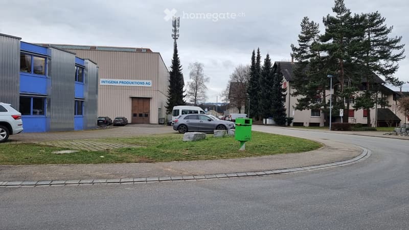 Industrial building named Intigena Produktion AG, with several cars parked in the area, including a gray car in front of the entrance, a white van, and a white SUV.