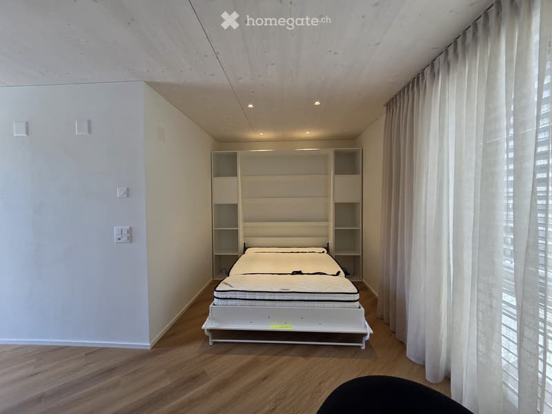 An empty room with a white bed, white shelves, and white walls. The bed is folded up against the wall. The room has a wooden floor and white curtains.