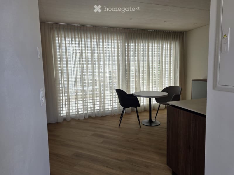 Empty room with wooden floor, white walls, and large white curtains. A round table with two chairs is placed near the window. A wooden cabinet is on the right side.