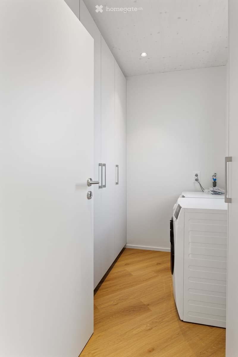 A modern laundry room with white walls, wooden floor, and built-in cabinets. It features a white washing machine and dryer, and a power outlet on the wall.
