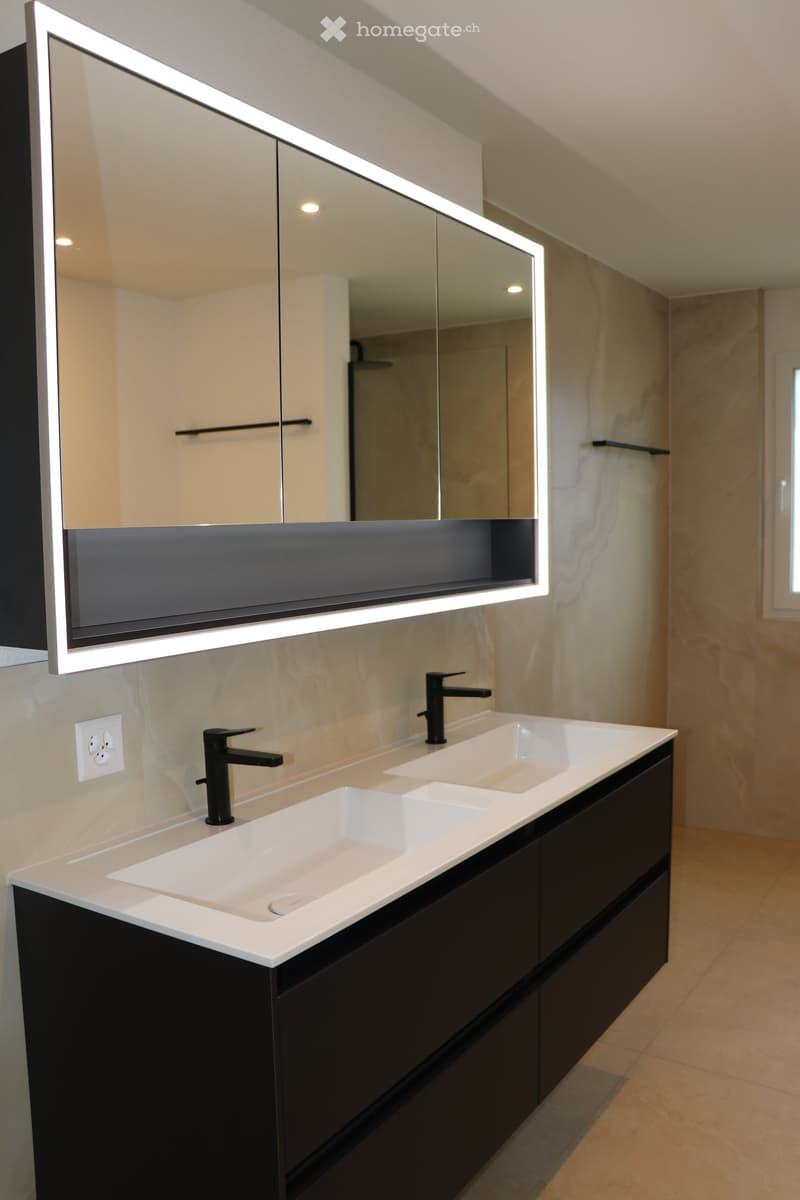 modern bathroom with double sink vanity, black cabinet, large mirror with integrated lighting, white tiled floor, silver faucets, power outlet