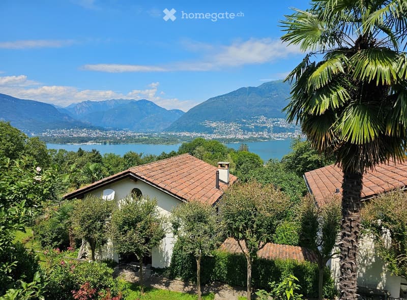 Traditional house with brown roof tiles, chimney, nestled among trees with lake and mountains in the background