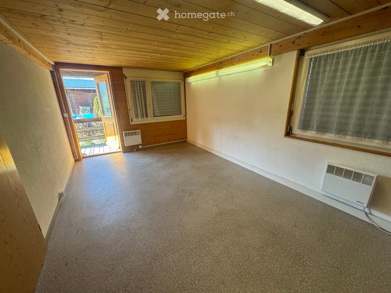 Empty room with wooden ceiling, carpeted floor, and large windows with curtains. Two wall-mounted heating units are visible, and there is an open doorway leading outside.