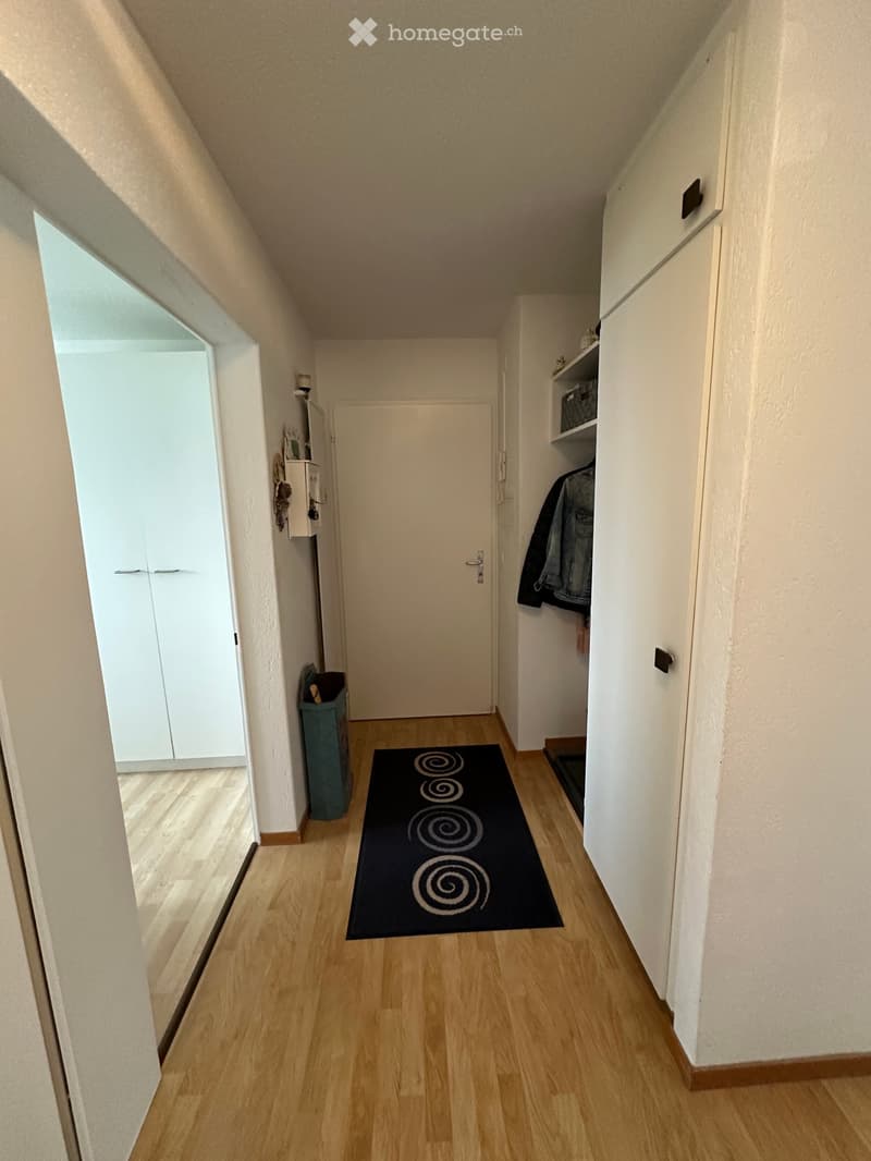 Corridor with wooden floors, two white doors, one open leading to another room, a trash bin, a black patterned rug, and a closet with a basket.