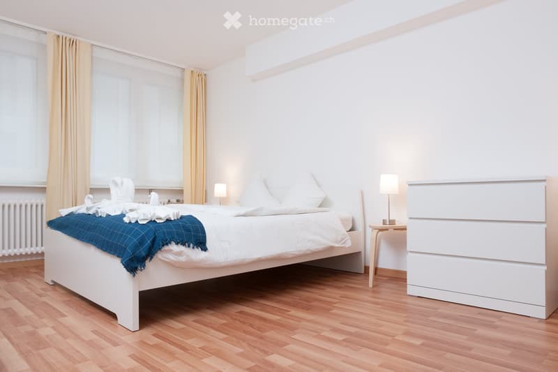 Modern styled bedroom, white bed with blue blanket, two white nightstands with lamps, yellow curtains, wooden parquet flooring, white wardrobe with drawers, radiator