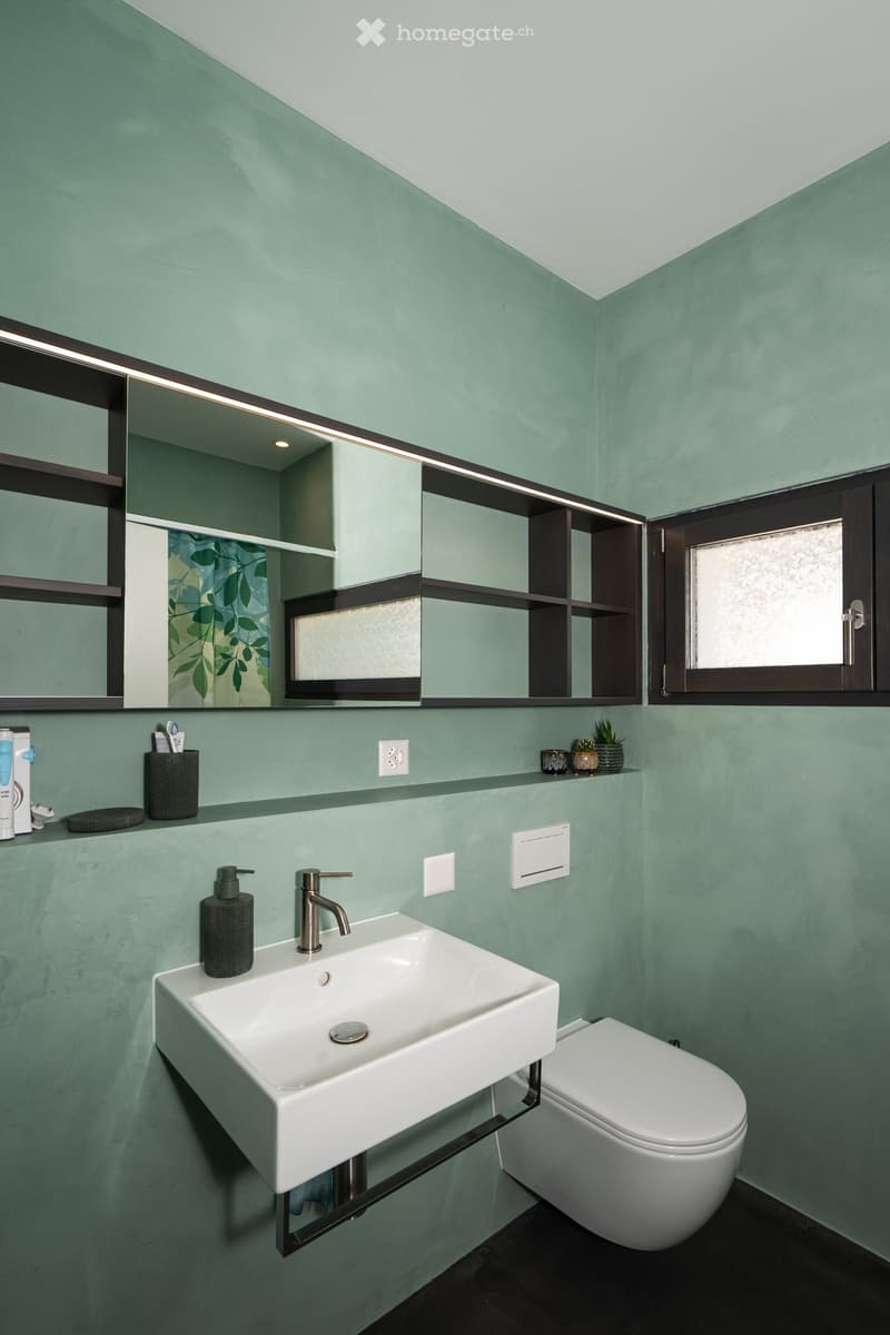 Bathroom with green walls, white sink, white toilet, mirror, shelves, and a window