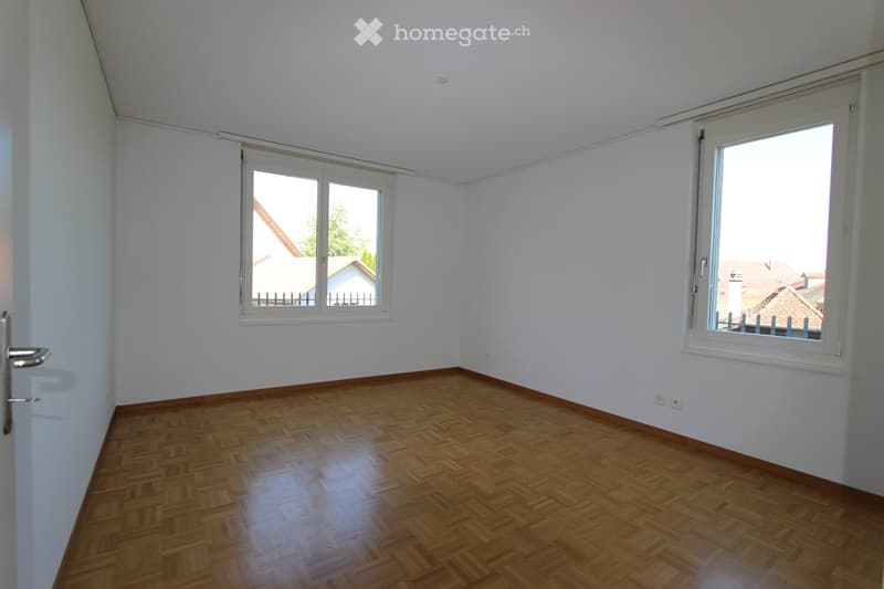 A spacious empty room with wooden floors, white walls, and large windows that allow natural light to enter.